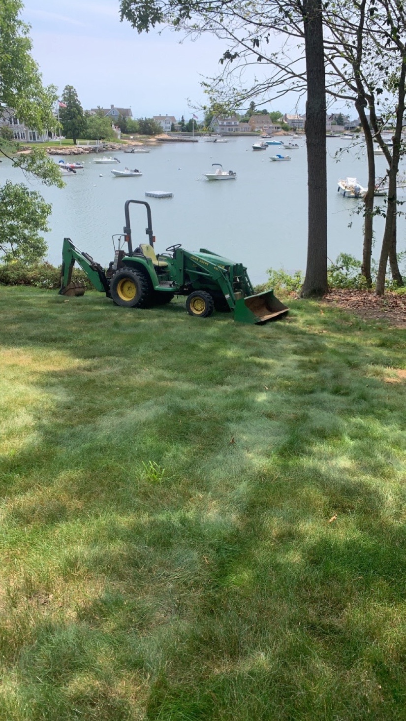 Compact tractor with backhoe attachment on a waterfront lawn overlooking moored sailboats